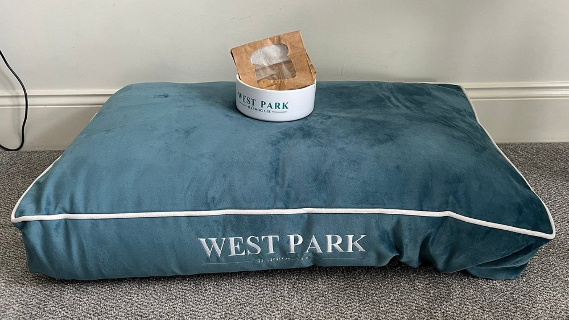 A plush teal dog bed and matching white pet bowl, both branded with The West Park Hotel Harrogate logo, featuring a bag of dog treats.