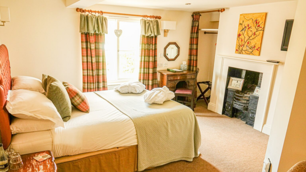 A rustic bedroom at The Oak Tree Inn in Helperby, featuring a large bed with white linens and green cushions, tartan curtains, a vintage wooden desk and a traditional black iron fireplace.