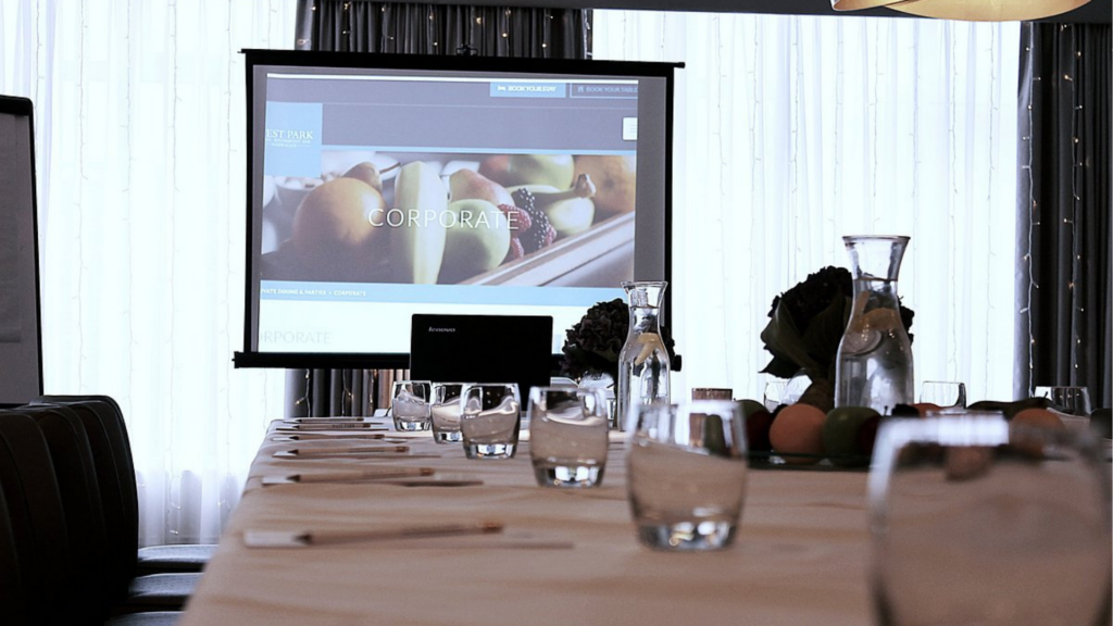 A conference table set with notebooks and glasses in front of a projector screen displaying a corporate presentation.