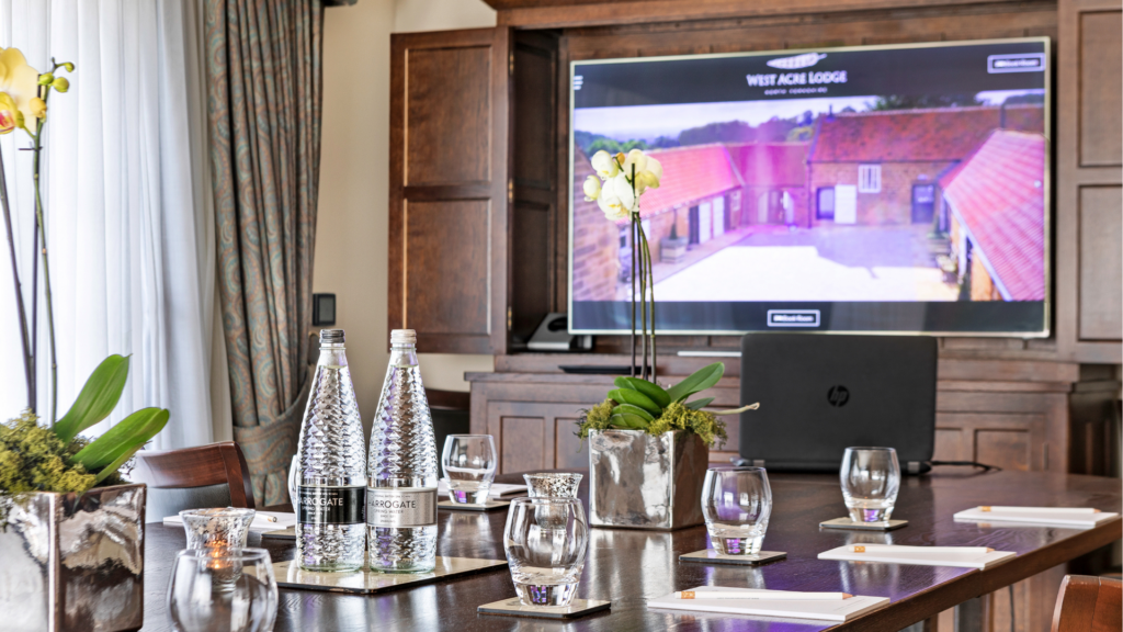 A boardroom table arranged with bottled spring water, glasses and note pads, featuring a large display screen in the background.